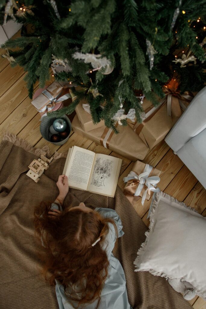 Teen girl reading under a Christmas tree, surrounded by gifts and decorations.